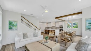 A spacious living area with a white sofa, a coffee table, and a dining area with wooden chairs and a table. A staircase with a wooden railing leads to an upper level.