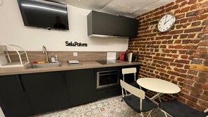 A compact kitchen with a built-in oven, a sink, and a small dining area with a table and two chairs. A clock is mounted on the brick wall, and a flat-screen TV is visible above the counter. - Quartier Voltaire - Rue Pache (Paris)