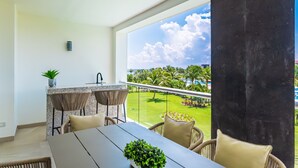 Un balcon offrant une vue sur un jardin luxuriant et des palmiers, un coin repas avec des meubles en osier, et une cuisine dotée d’un plan de travail en granit.