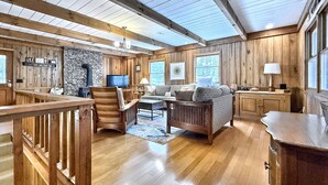 A spacious cabin interior with a wooden floor, a stone fireplace, a sofa set, a dining table, and a kitchen area. - Creekside,firepit,wood stove,family&dog friendly,covered deck dining (Scaly Mountain)
