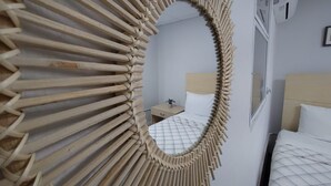 A room with a unique wooden sunburst mirror, a bed with a white quilt, and a small bedside table with a plant.