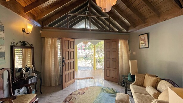 A spacious room with a high ceiling, a wooden door leading outside, a sofa, a chair, and a table with a lamp. - Yancey Largo Villa Perfect Jamaica Ironshore Getaway w Private Pool and Onsite Staff Services by RedAwning (Montego Bay)