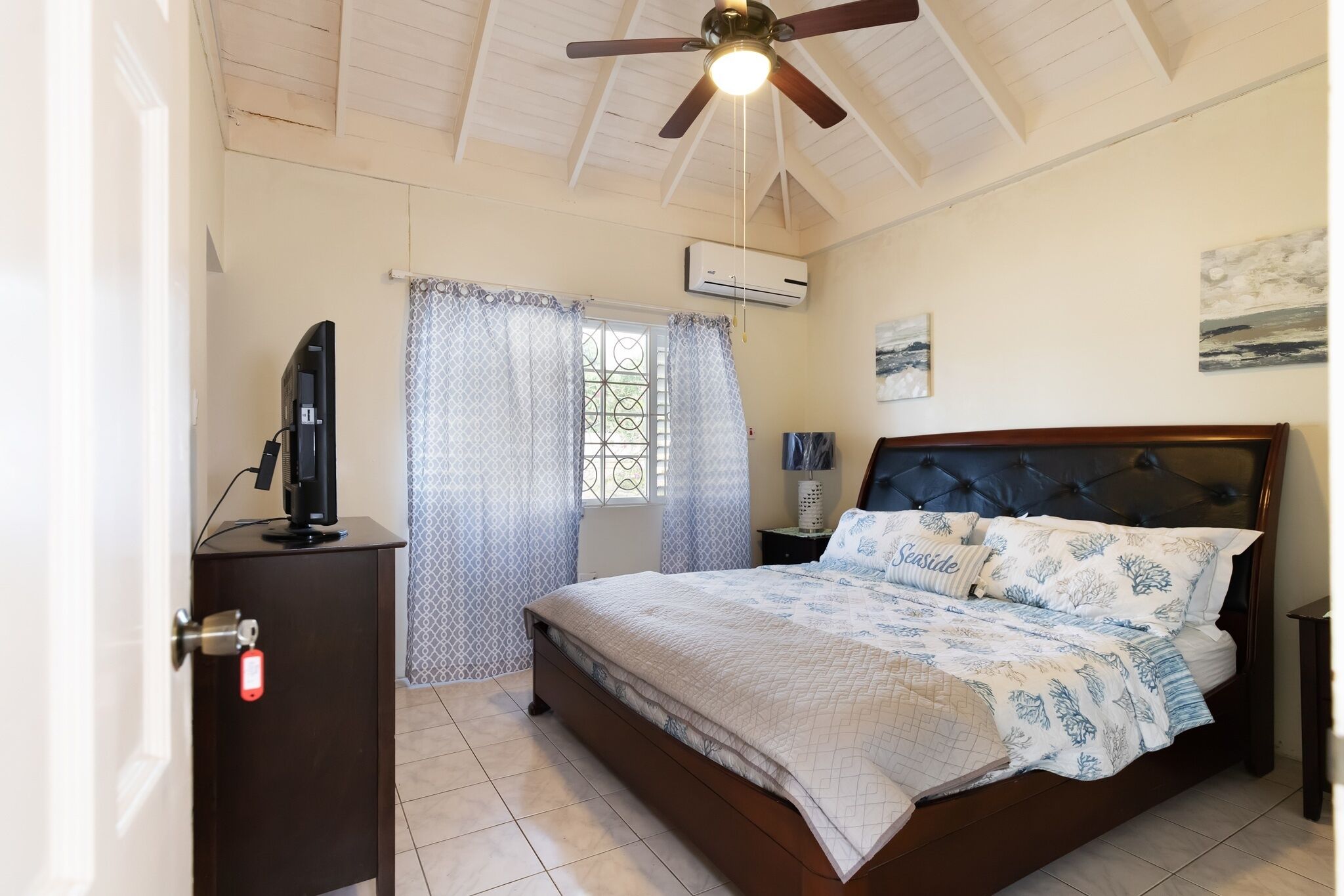 Een slaapkamer met een bed, een plafondventilator, een televisie op een kast en een airconditioner.