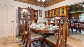 A dining area with a wooden table set for six, surrounded by wooden chairs. A wooden cabinet with glass doors is to the left, and a kitchen area is visible in the background. - Yancey Largo Villa Perfect Jamaica Ironshore Getaway w Private Pool and Onsite Staff Services by RedAwning (Montego Bay)