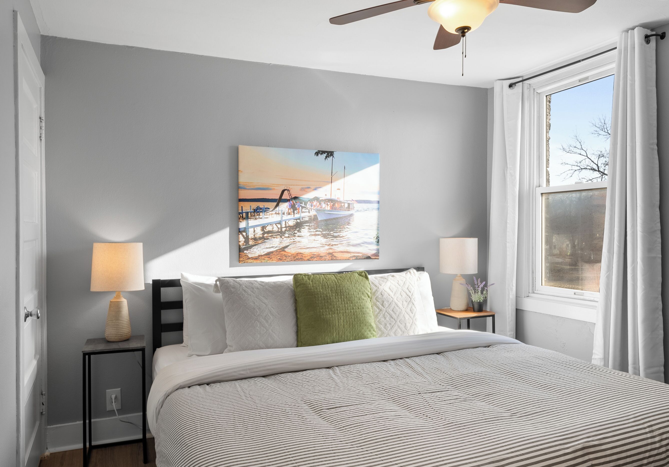 A bedroom with a bed, two bedside tables with lamps, a painting on the wall, and a window with curtains.