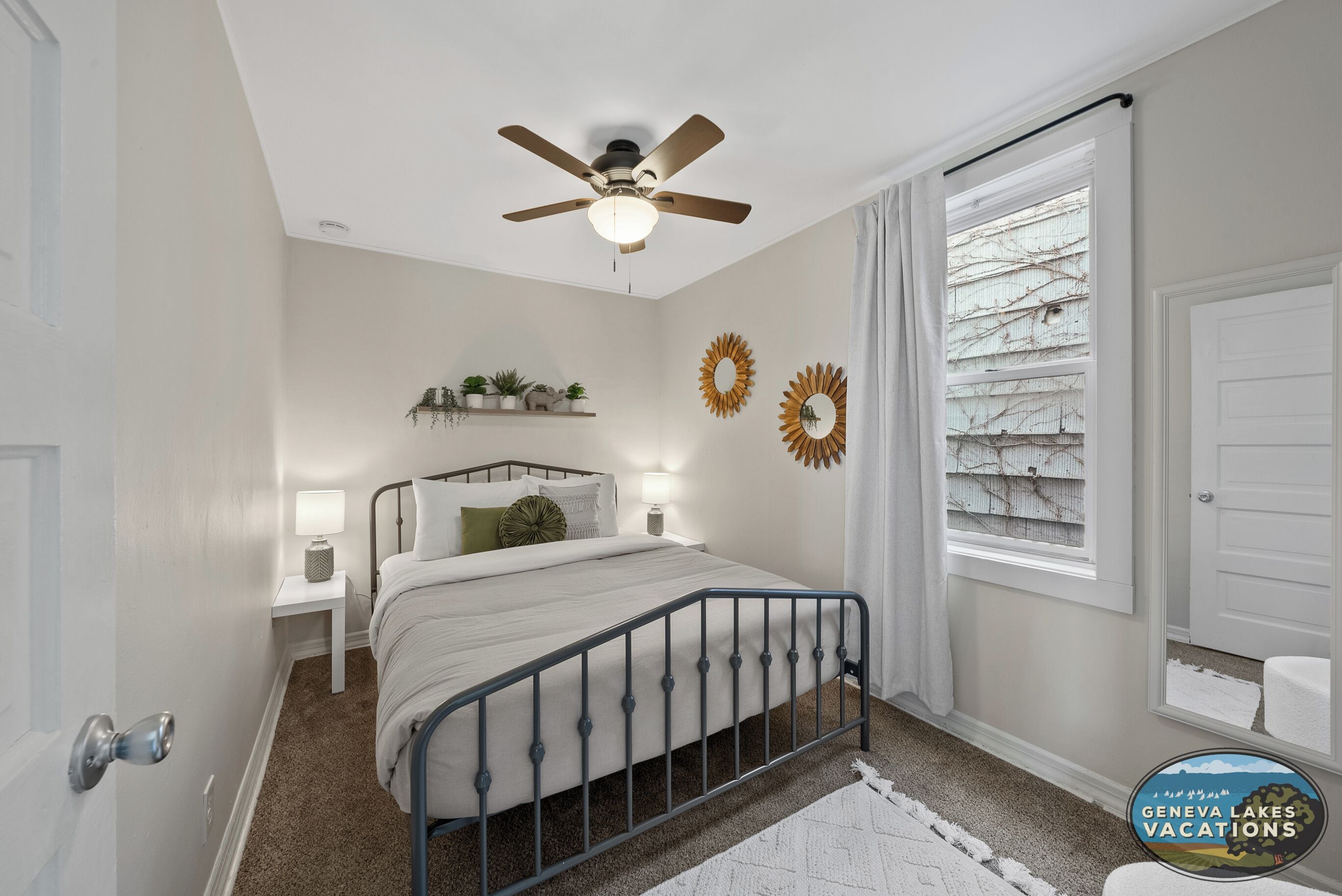 Un dormitorio con cama, mesitas de noche, ventilador de techo, ventana con cortinas y adornos en las paredes.