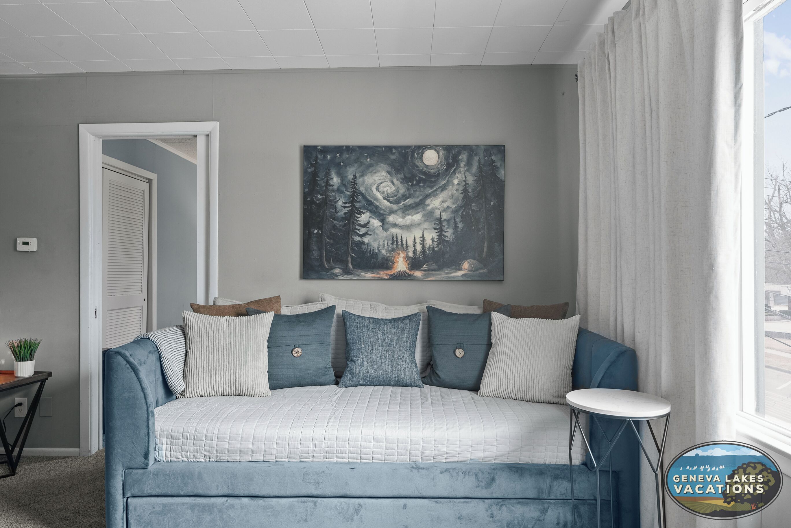 A bedroom with a bed, a night sky painting, a window with curtains, and a small table.