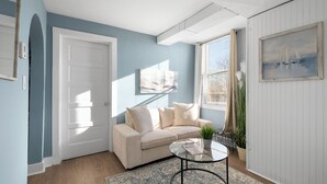 A living room with a beige sofa, a glass coffee table, a patterned rug, a painting on the wall, and a potted plant. - Broad Street Suites 1,2 & 3 |Sleeps 20 |Parking for 7 Vehicles |Downtown |Events |Horticultural Hall (Lake Geneva)