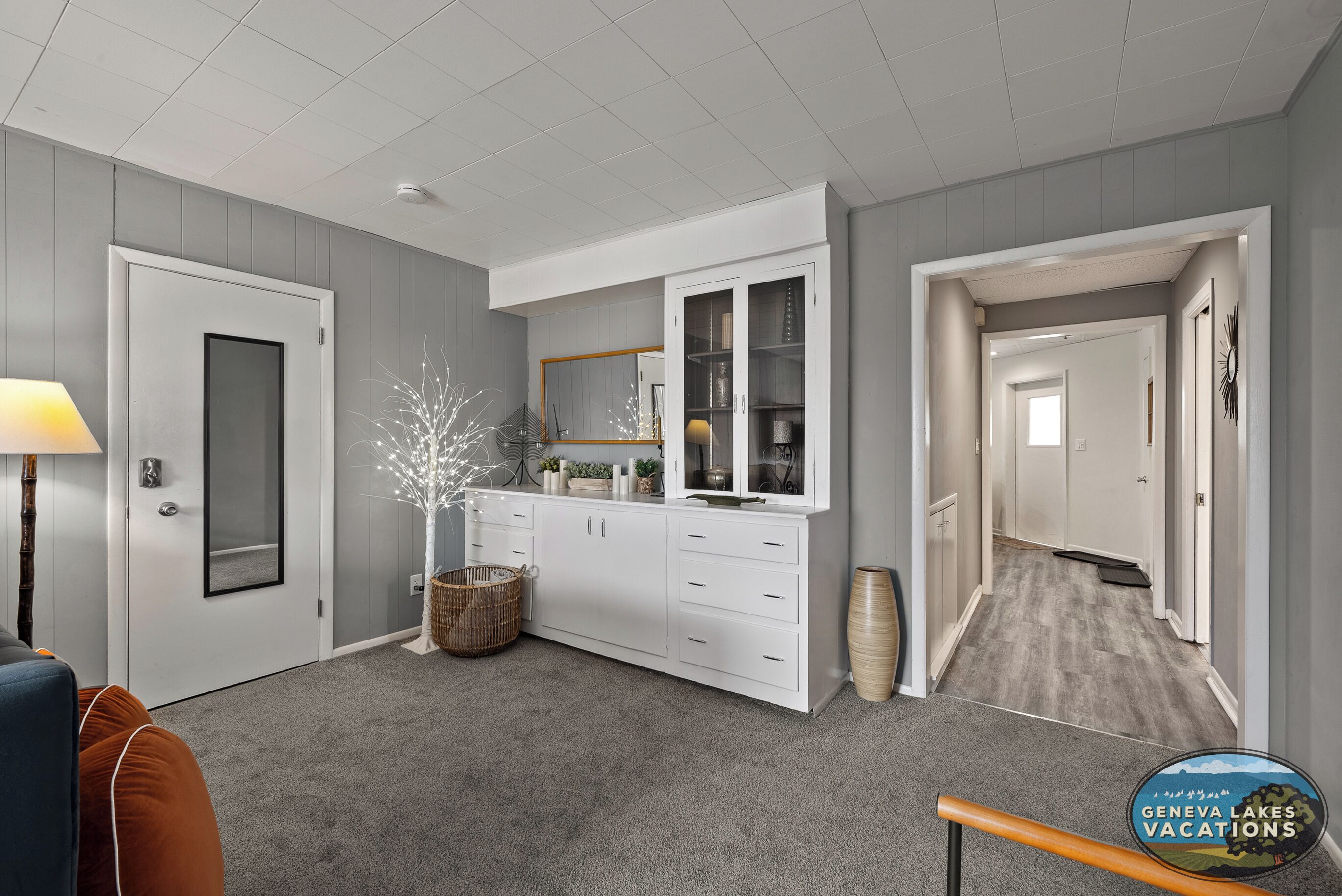 Un interior moderno con alfombra gris, muebles blancos, una cesta de mimbre y un jarrón grande.