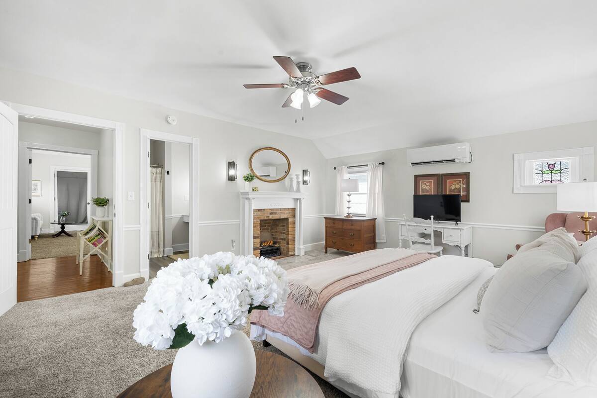 A bedroom with a bed, a desk, a TV, a fireplace, and a ceiling fan.