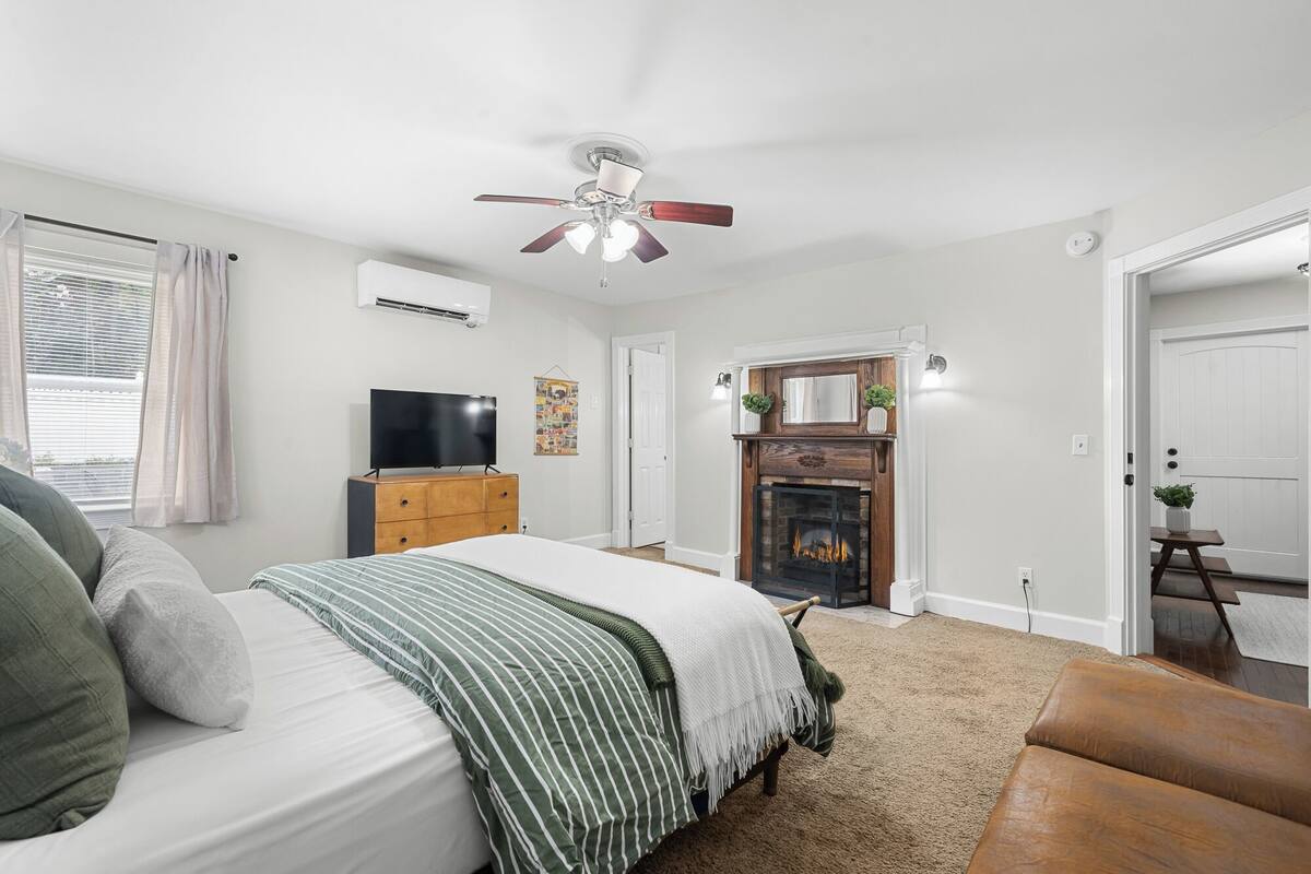 A bedroom with a bed, a television, a ceiling fan, a fireplace, and a door leading to another room.