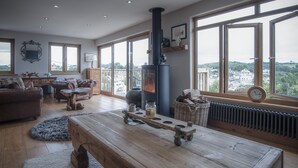 A spacious living room with a large wooden table, a fireplace, and large windows offering a view of the city. - Bayview House - 4 Bedroom - Saundersfoot (Saundersfoot)