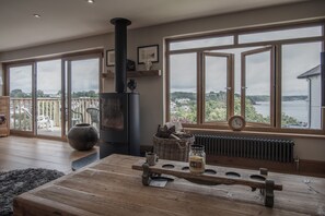 A spacious room with large windows offering a view of the sea, a wood-burning stove, and a rustic wooden table with a jar of jam.