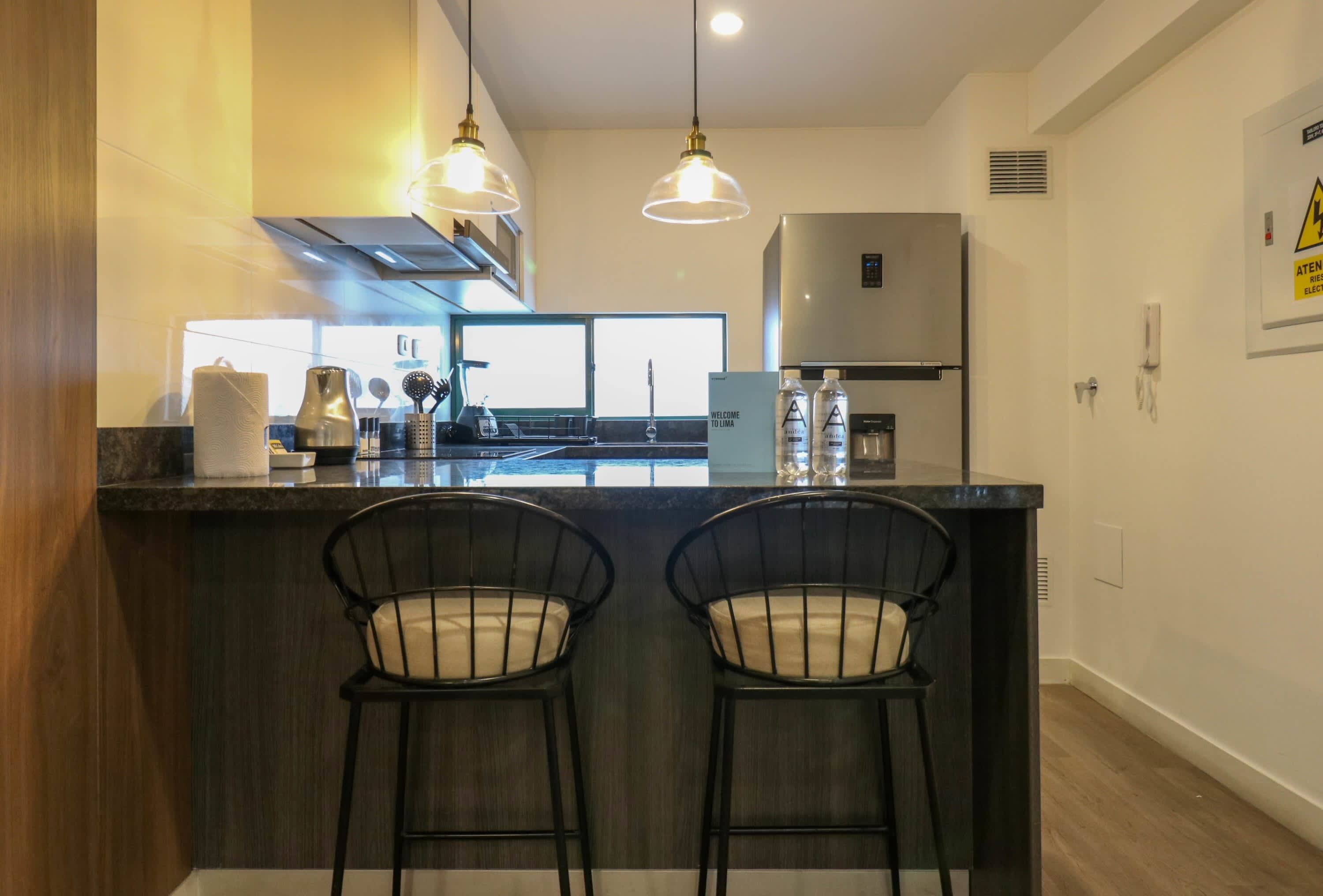 A modern kitchen with a central island, two bar stools, pendant lighting, a stainless steel refrigerator, and a view of the outside.