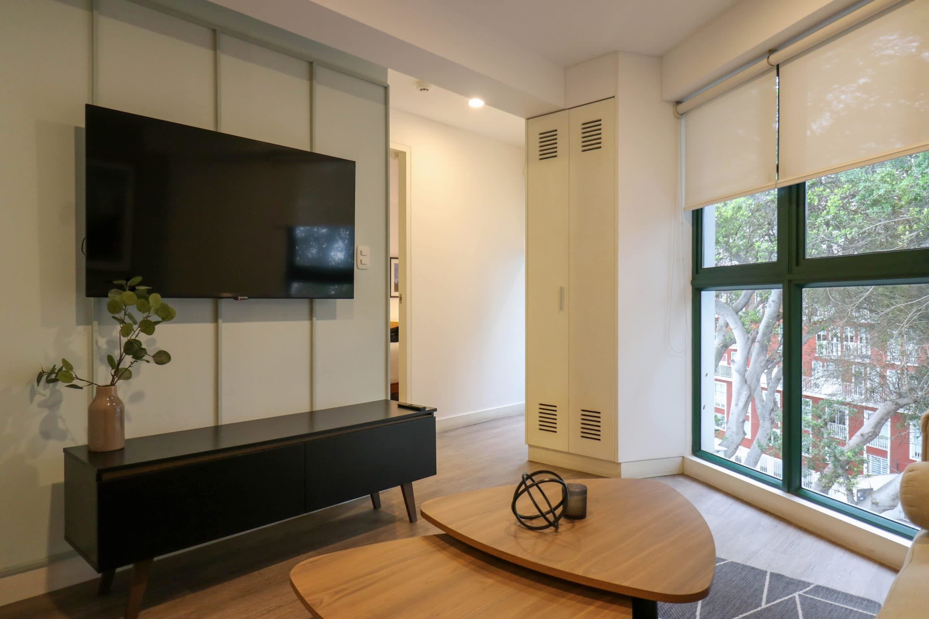 A modern living room with a flat-screen TV, a dark console table, a wooden coffee table, and a large window offering a view of the outside.