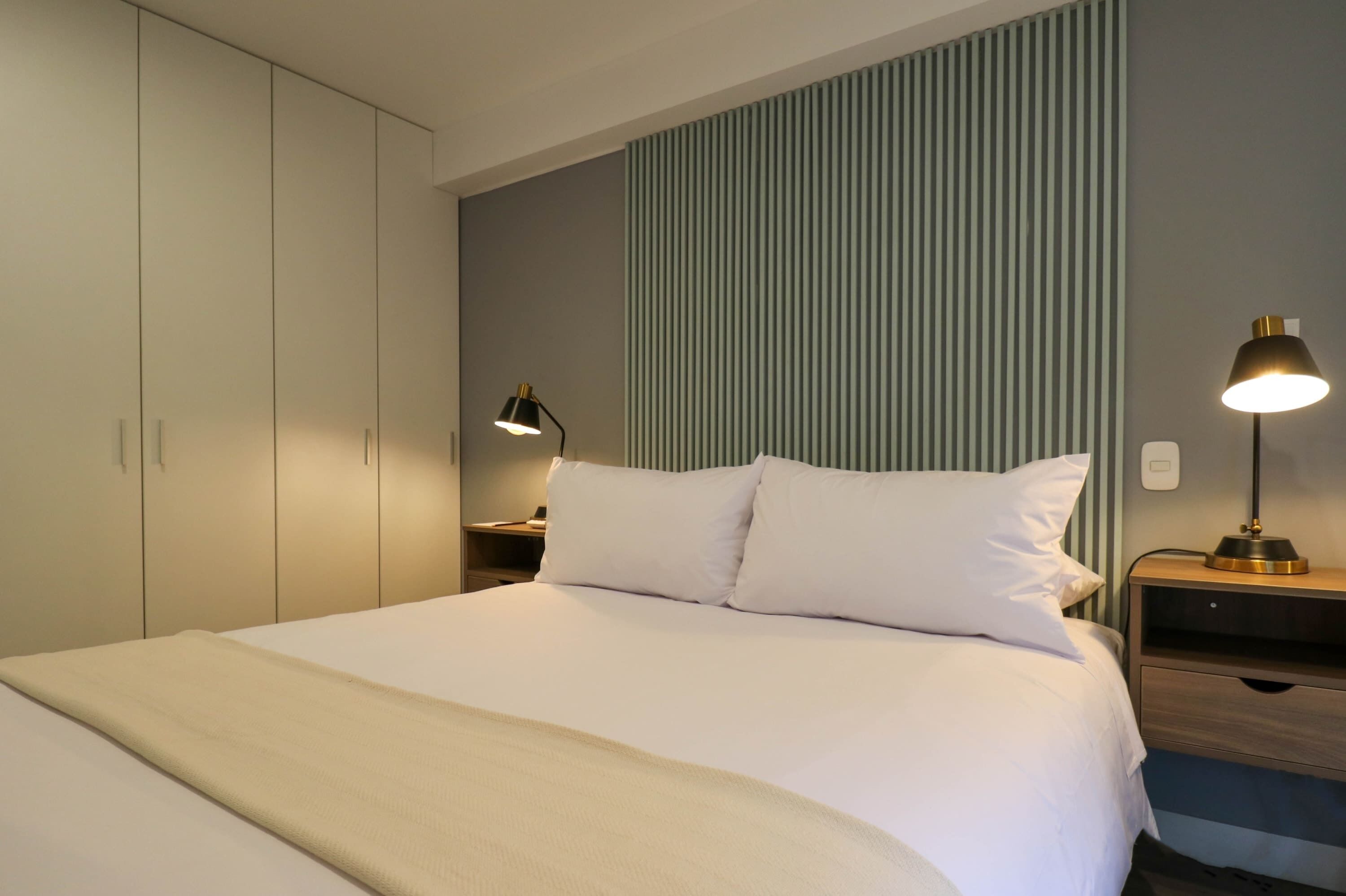 A neatly made bed with white linens, a bedside table with a lamp, and a wardrobe.