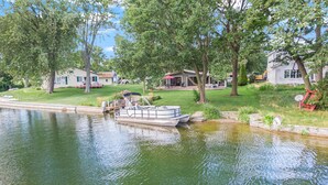 A houseboat docked at a calm river, surrounded by lush greenery and residential buildings.