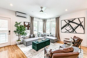 Un salon moderne avec un canapé d’angle, un pouf vert, des fauteuils en cuir et un grand tableau abstrait.