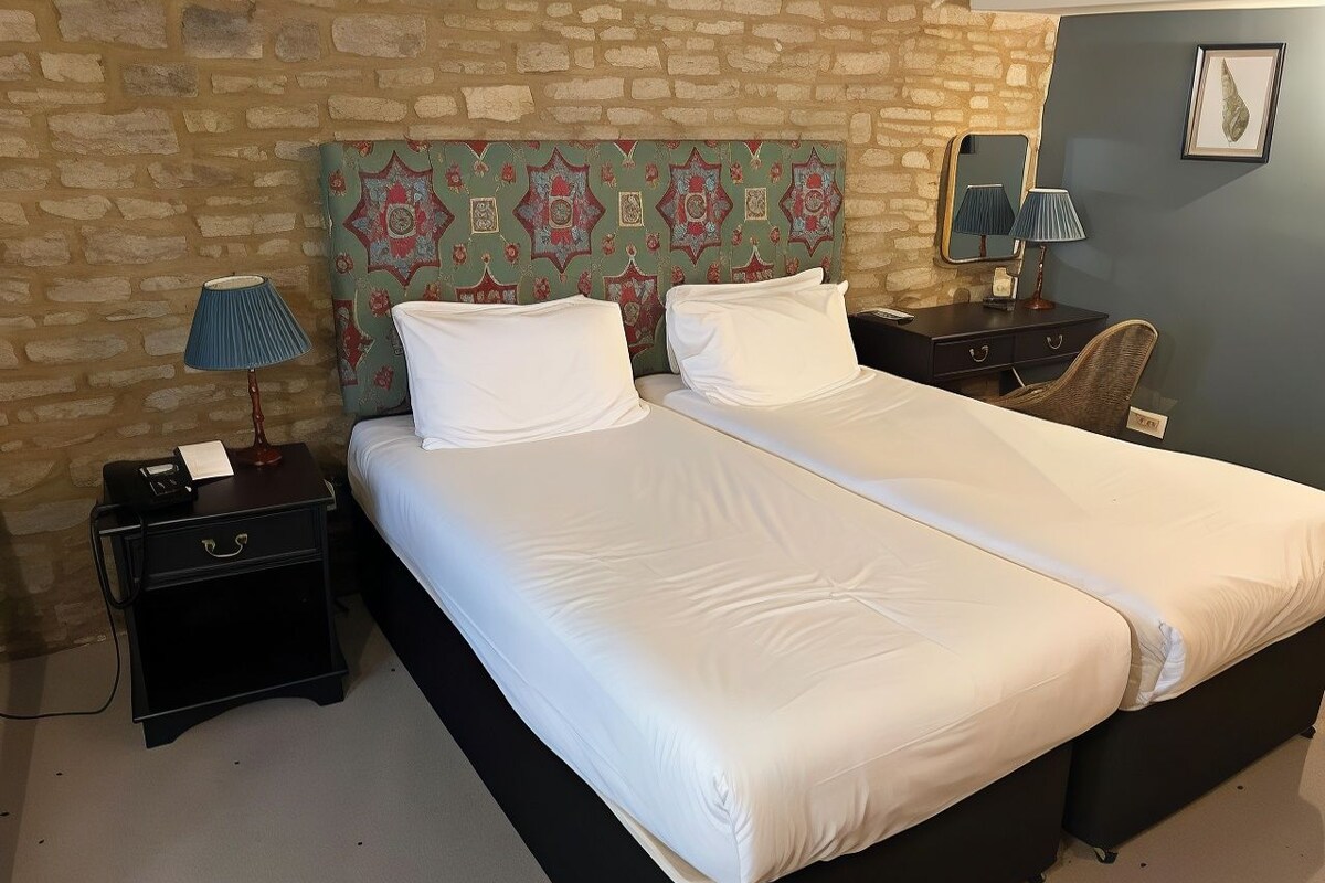 A double bed with white linens, a headboard with a floral pattern, a nightstand with a lamp, and a wall-mounted mirror.