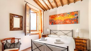 Une chambre à coucher avec un lit, une chaise en bois, une table de chevet, une lampe, un miroir et un grand tableau abstrait accroché au mur.