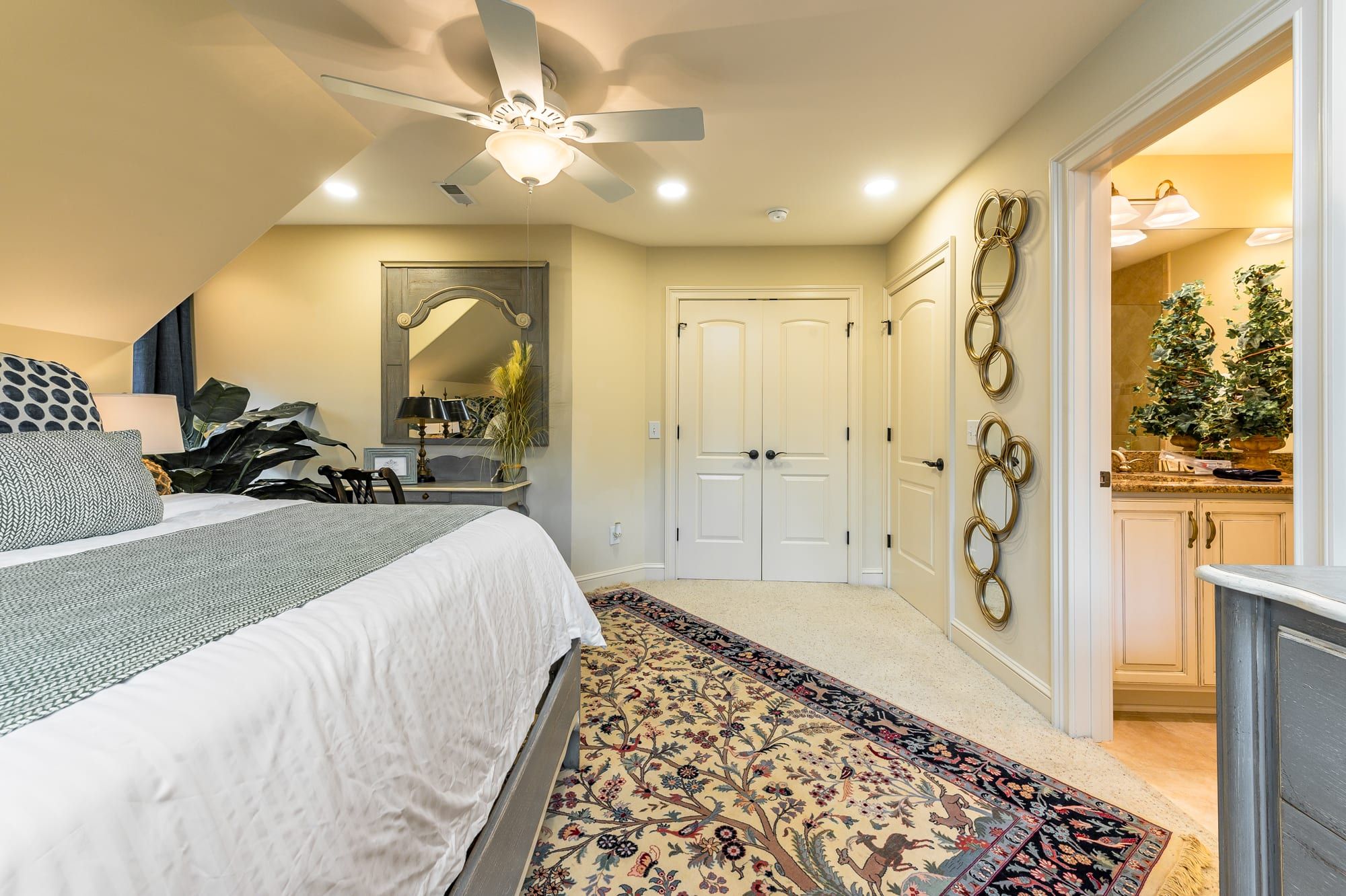 Une chambre à coucher comprenant un lit, un ventilateur de plafond, un miroir et une porte donnant sur une salle de bain.