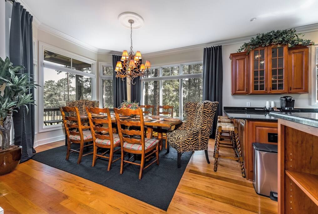 A dining area with a wooden table and chairs, a chandelier, and large windows with curtains.