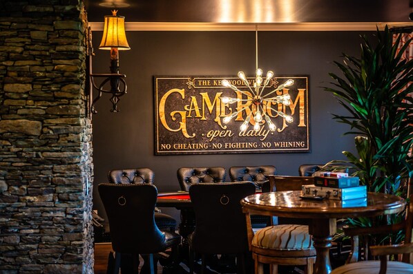 A cozy bar area with a stone wall, leather chairs, a wooden table, and a sign that reads "The CAMERUM open daily. No cheating. No fighting. No whining."