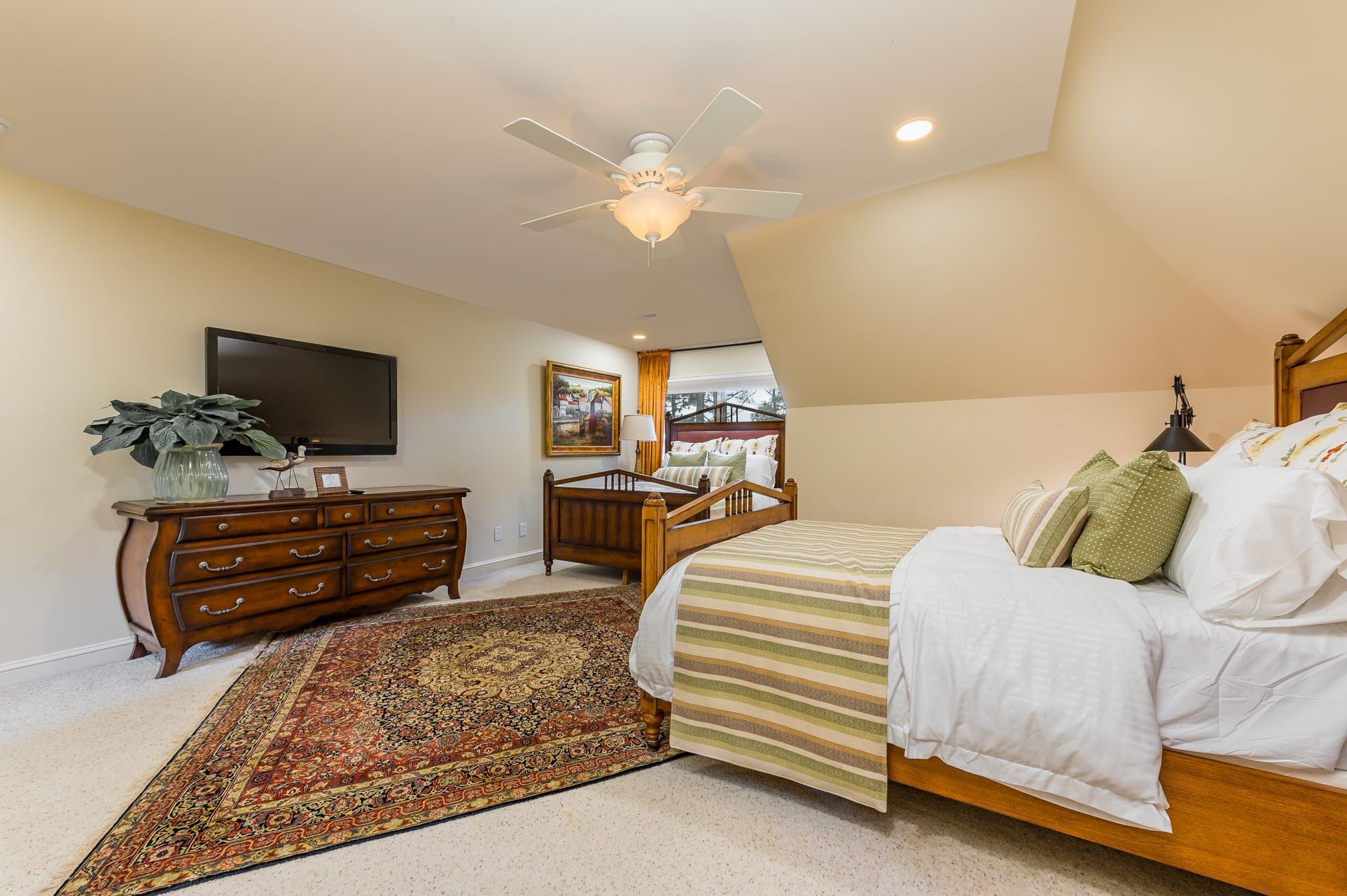 A bedroom with a bed, a wooden dresser, a TV, a ceiling fan, and a large window.