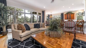Amplia sala de estar con mesa de centro de madera, sofá color beige con cojines estampados y un gran ventanal con vistas a árboles.