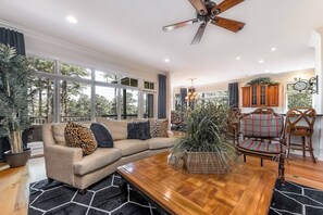 A spacious living room with a wooden coffee table, beige sofa with patterned cushions, and a large window offering a view of trees.