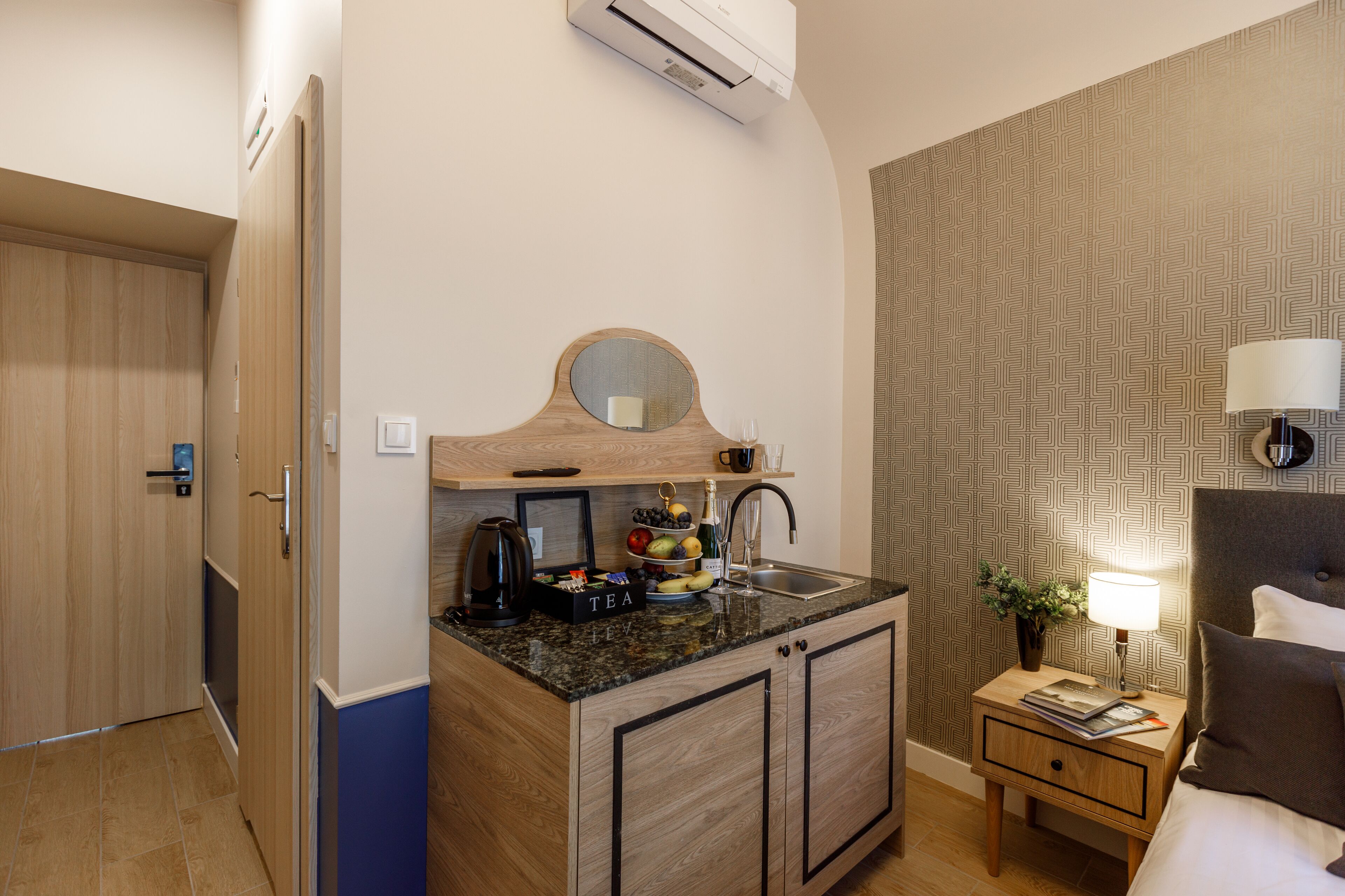 Habitación de hotel pequeña con kitchenette, una cama, una mesita de noche con lámpara y un papel tapiz estampado.