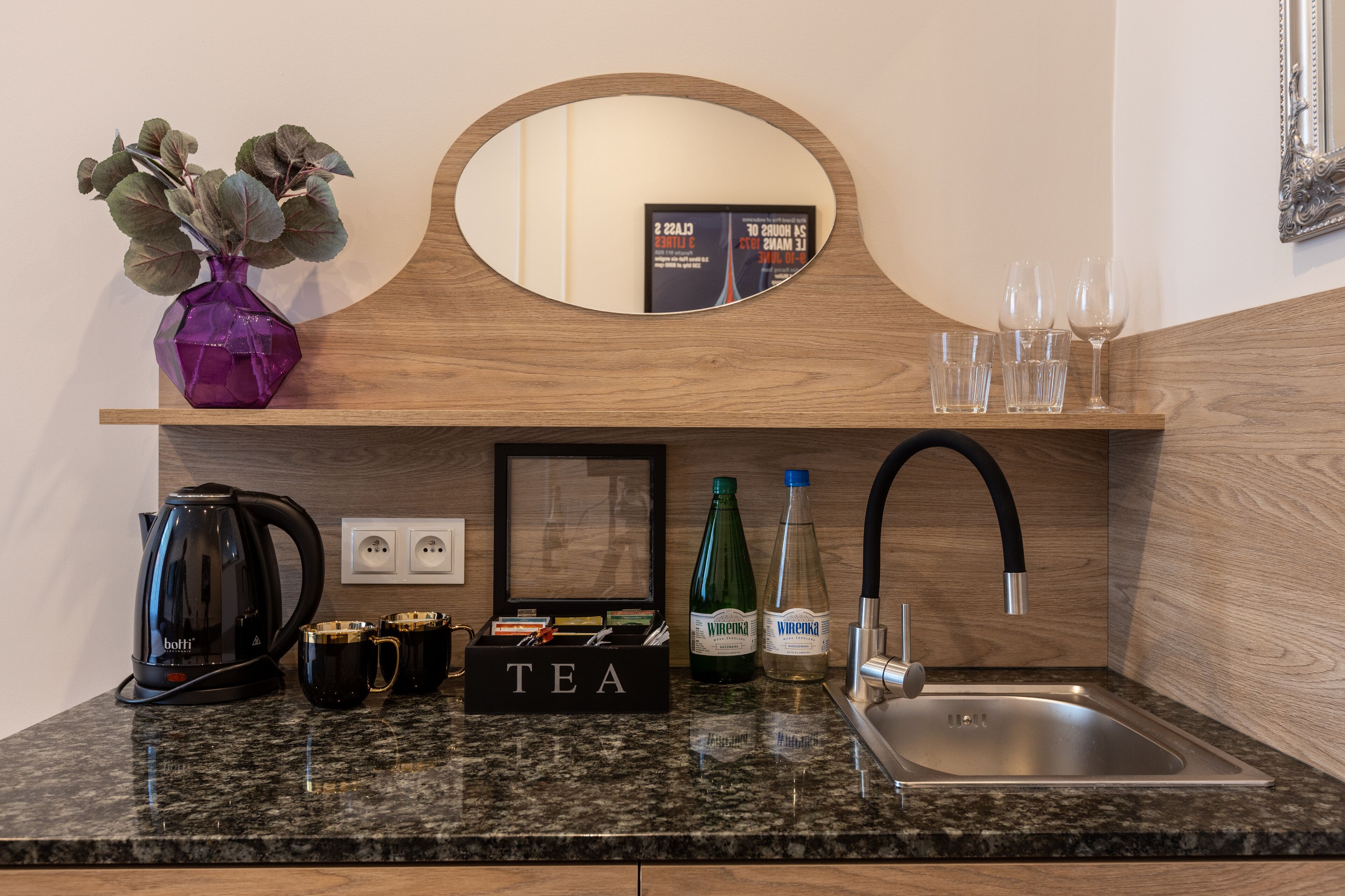 A kitchen counter with a coffee maker, tea box, bottles, glasses, and a mirror.