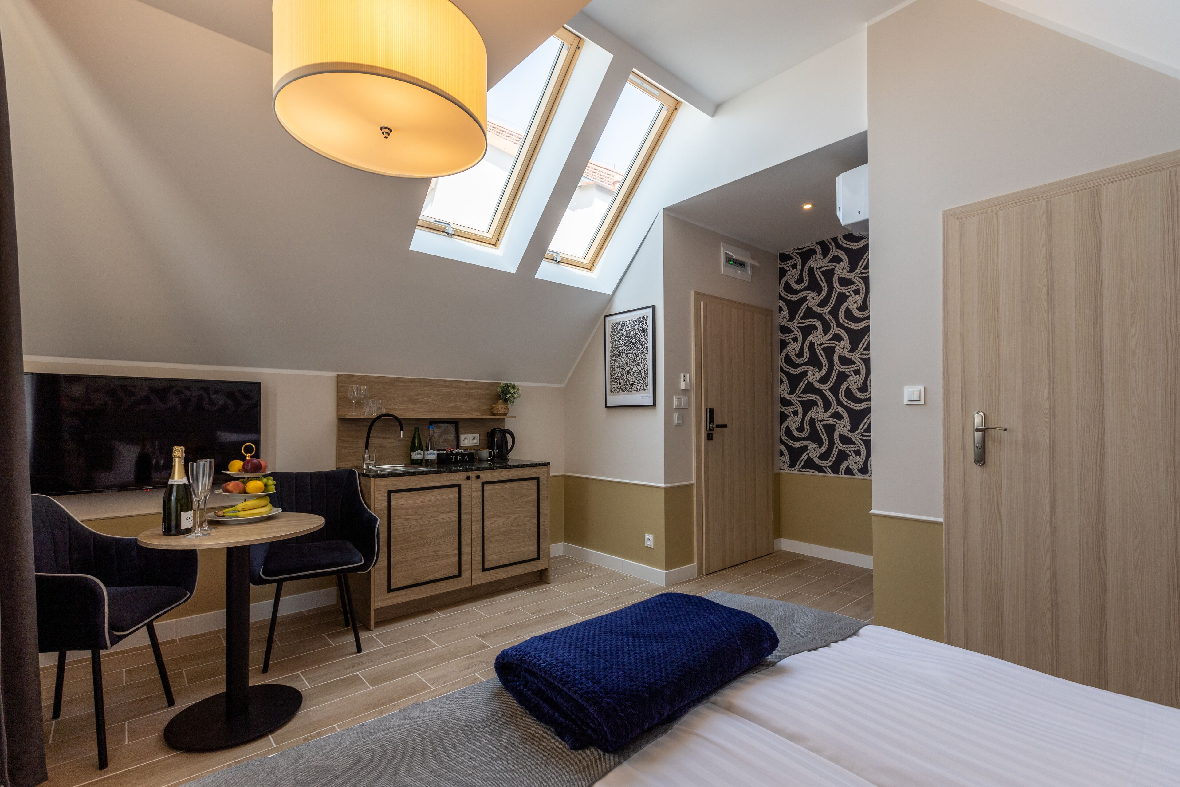 Una habitación compacta con cama, una mesa de comedor con sillas, kitchenette y un ventanal con tragaluces.