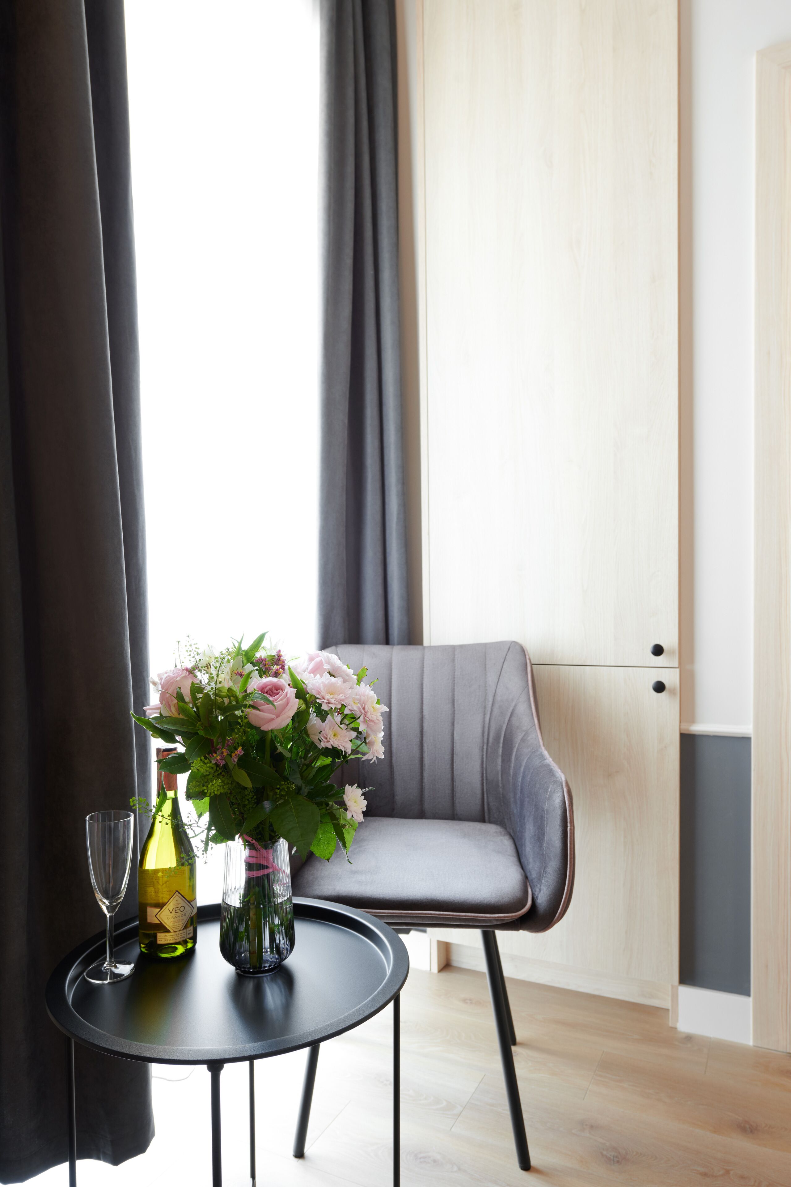 A modern room with a round black table, a grey upholstered chair, a vase with flowers, and a bottle of champagne.