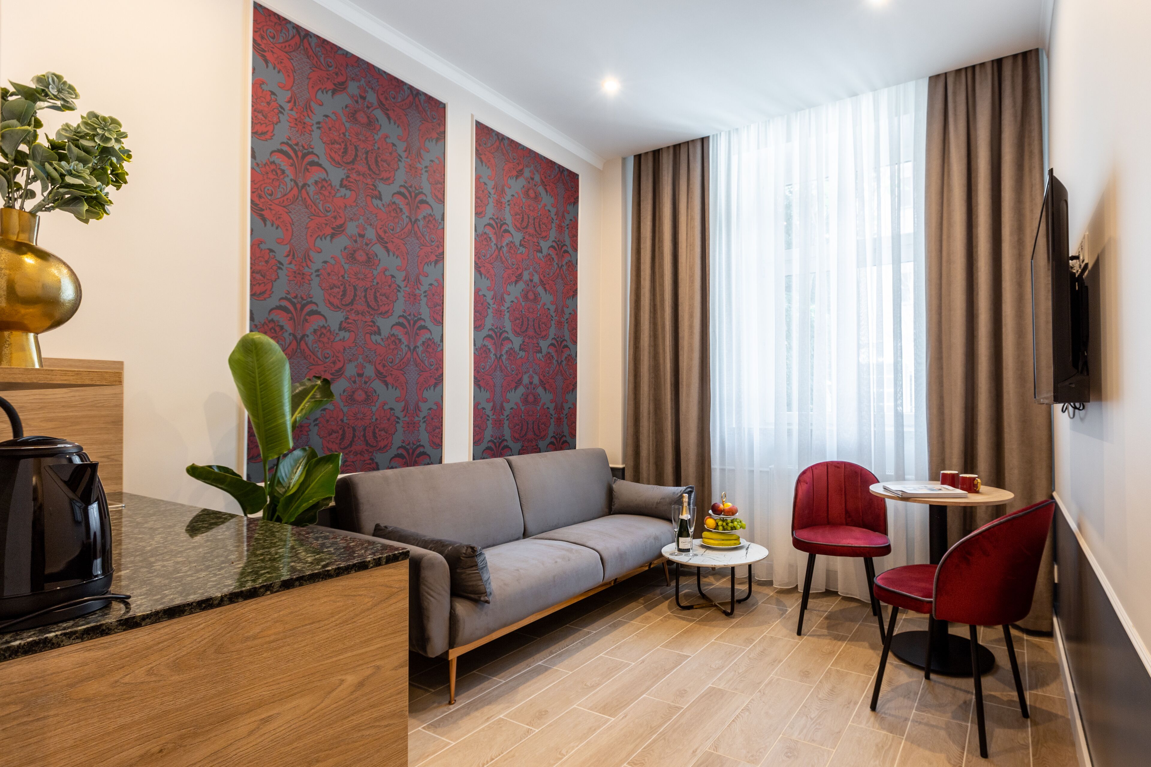 Un salon moderne avec un canapé, une petite table ronde et deux fauteuils rouges. On aperçoit une plante sur la gauche et un téléviseur à écran plat fixé au mur.