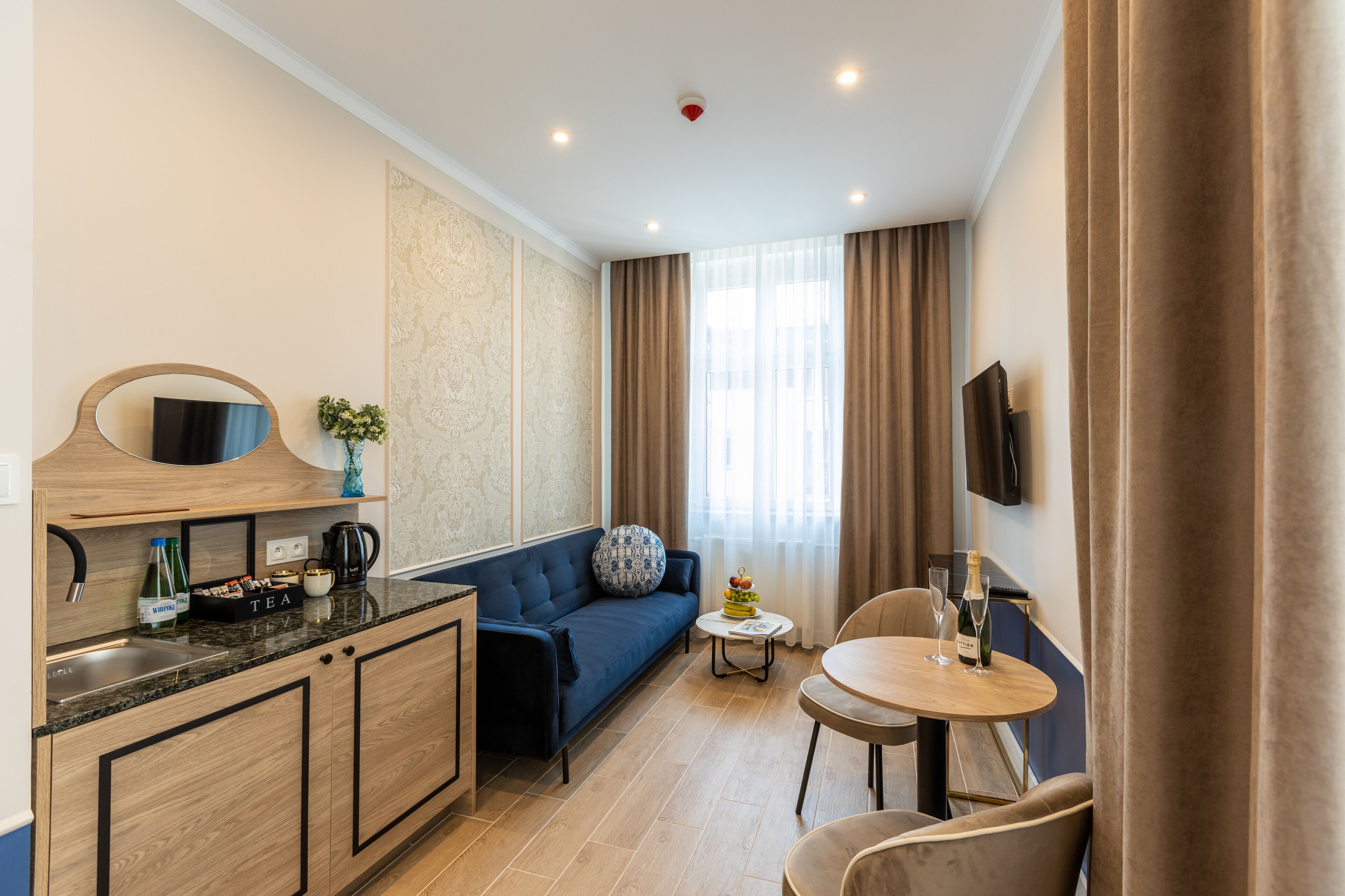 A modern hotel room with a kitchenette, a sofa, a small round table, and a TV mounted on the wall.