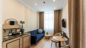 A modern hotel room with a kitchenette, a sofa, a small round table, and a TV mounted on the wall.