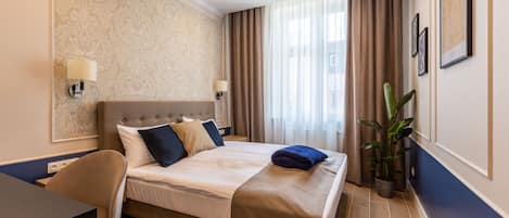 A hotel room with a bed, a desk, a chair, a potted plant, and a window with curtains.
