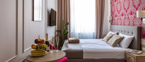 A hotel room with a bed, a bedside table, a chair, a small table with fruit and drinks, and a large window with curtains.