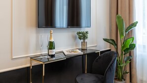 Quarto moderno com televisão de tela plana na parede, mesa de centro de vidro com uma garrafa de champanhe e copos, uma planta em vaso e uma poltrona confortável.