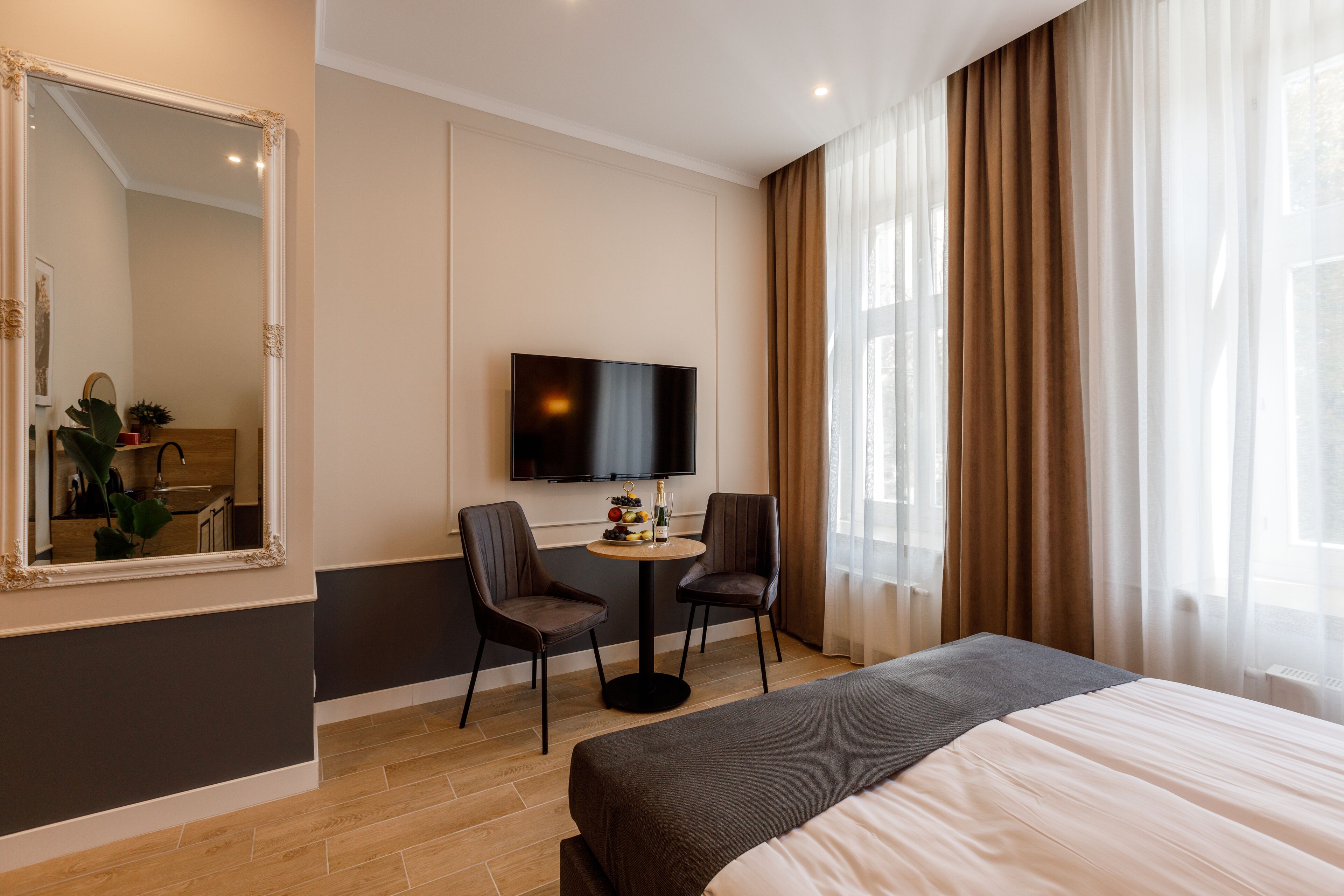A hotel room with a bed, a TV, two chairs, a small table with a bottle and fruit, and a large mirror.