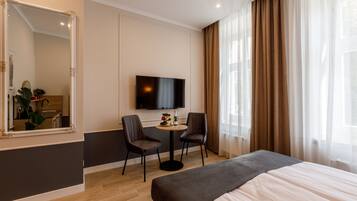 A hotel room with a bed, a TV, two chairs, a small table with a bottle and fruit, and a large mirror.