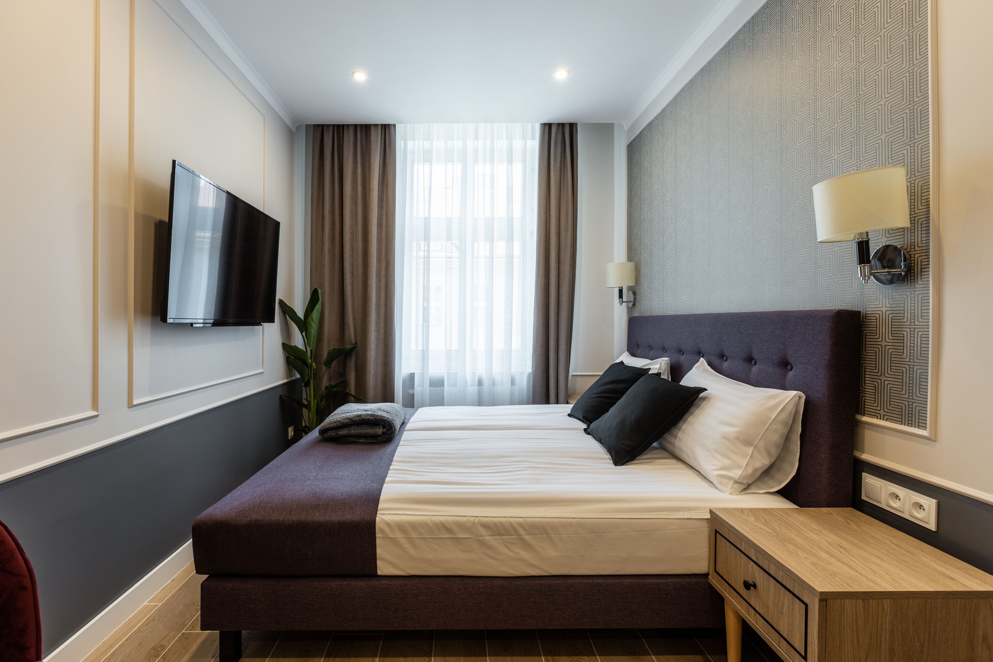 A hotel room with a large bed, a TV mounted on the wall, a bedside table with a lamp, and a window with curtains.