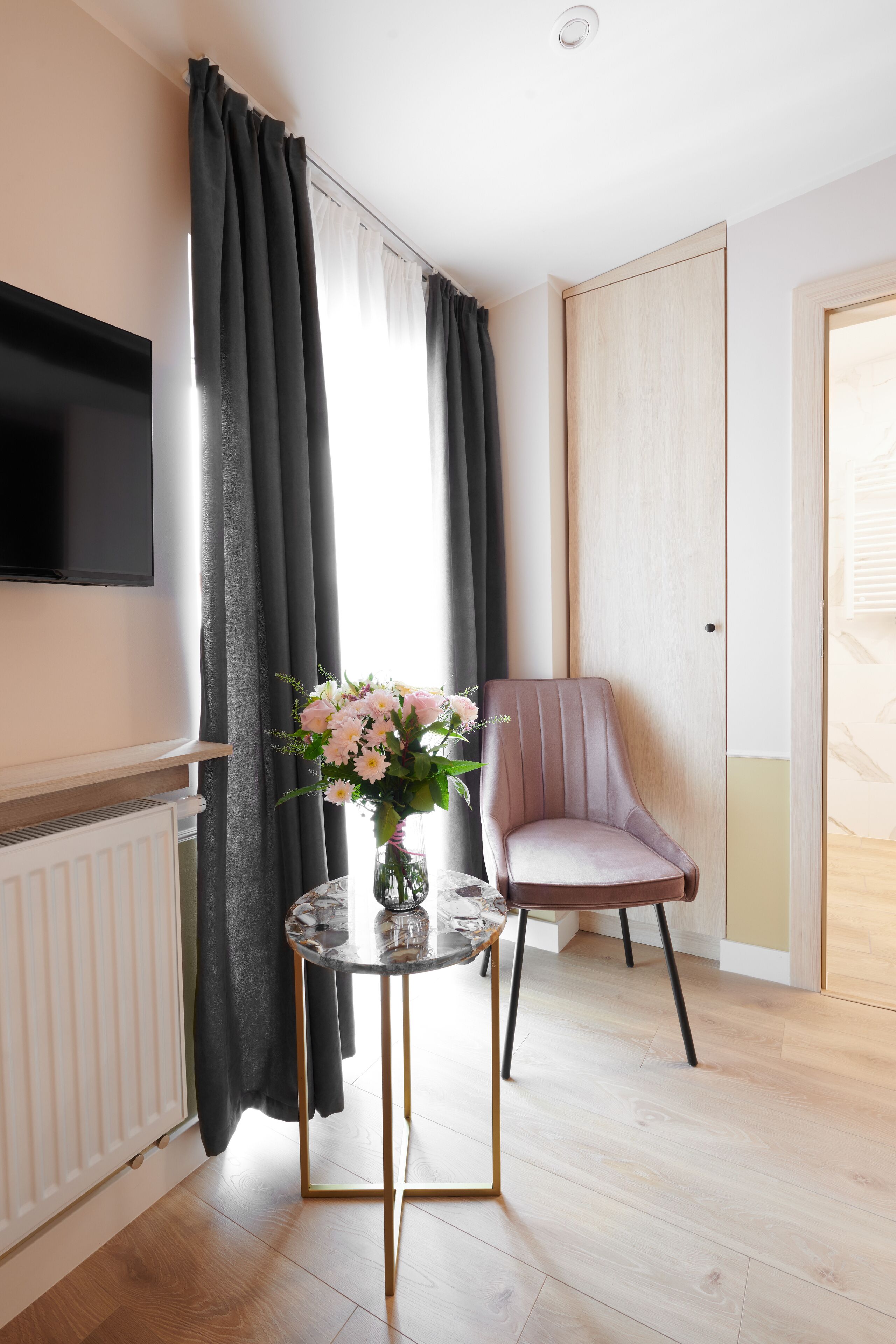 A modern living room with a flat-screen TV, a round table with a vase of flowers, a cushioned armchair, and a wooden floor.