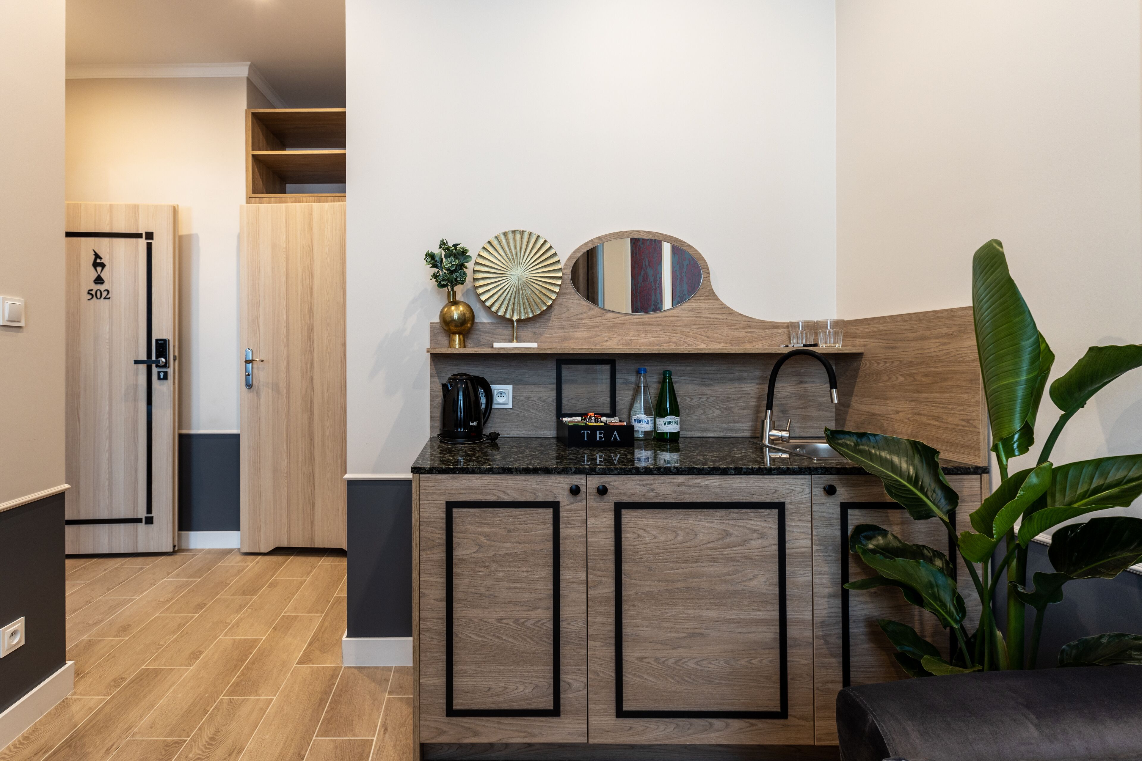 Una cocina moderna con armarios de madera, una encimera negra y una cafetera. Hay una planta en el lado derecho y una puerta con el número 502 a la izquierda.