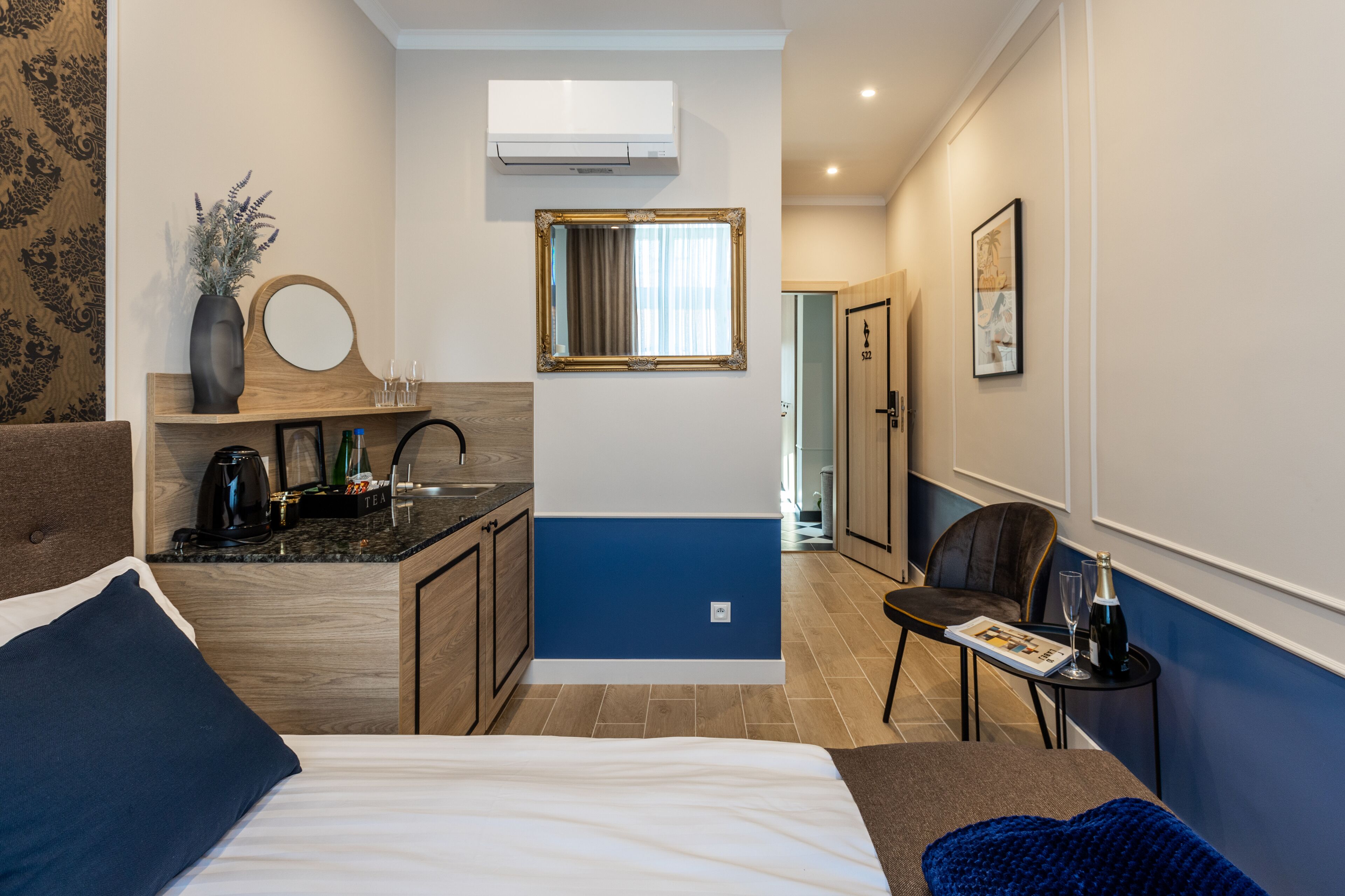 A compact hotel room with a bed, a small kitchenette, a sitting area, and a door leading to a hallway.