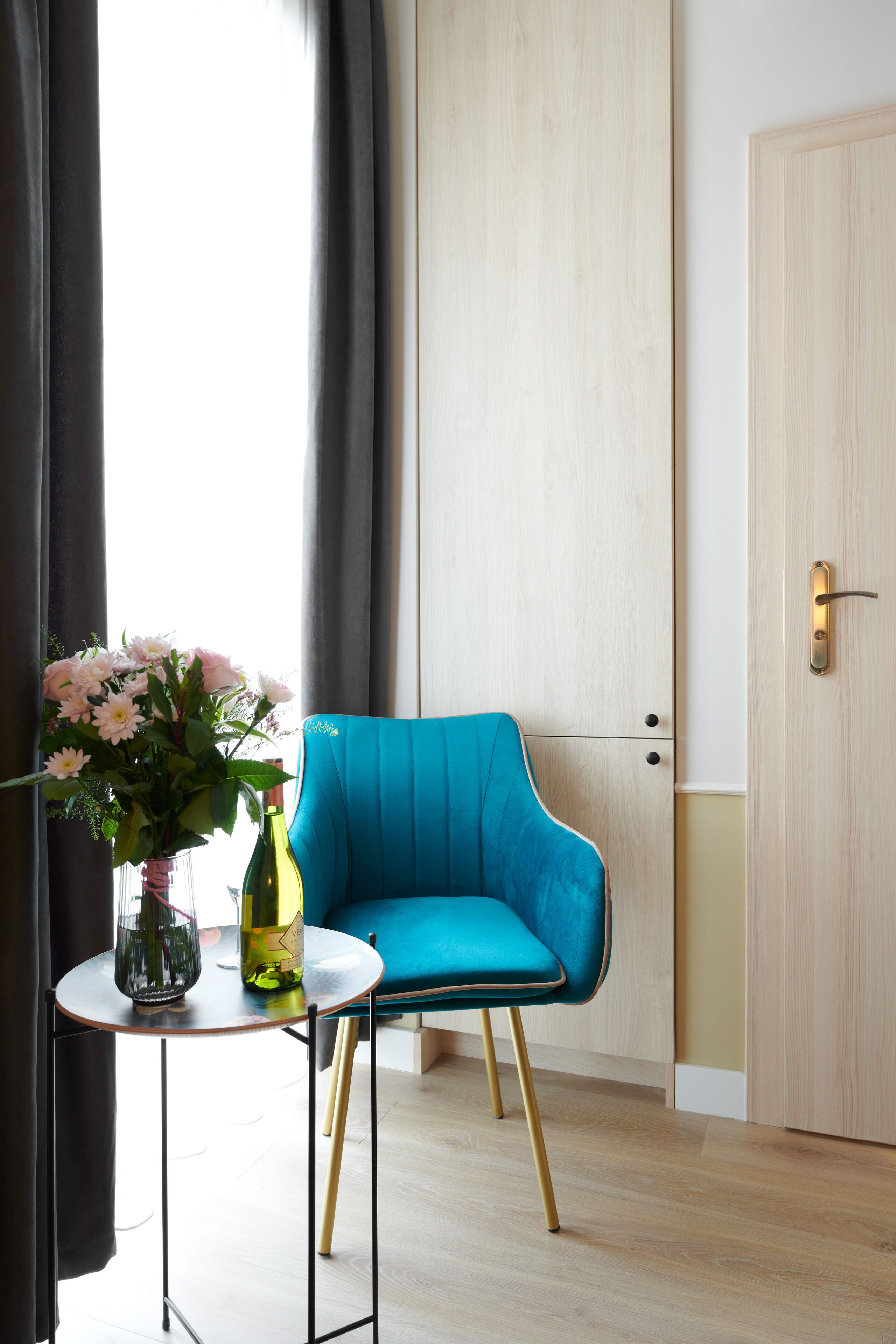A room with a teal chair, a small round table with a vase of flowers and a bottle, and a wooden door.