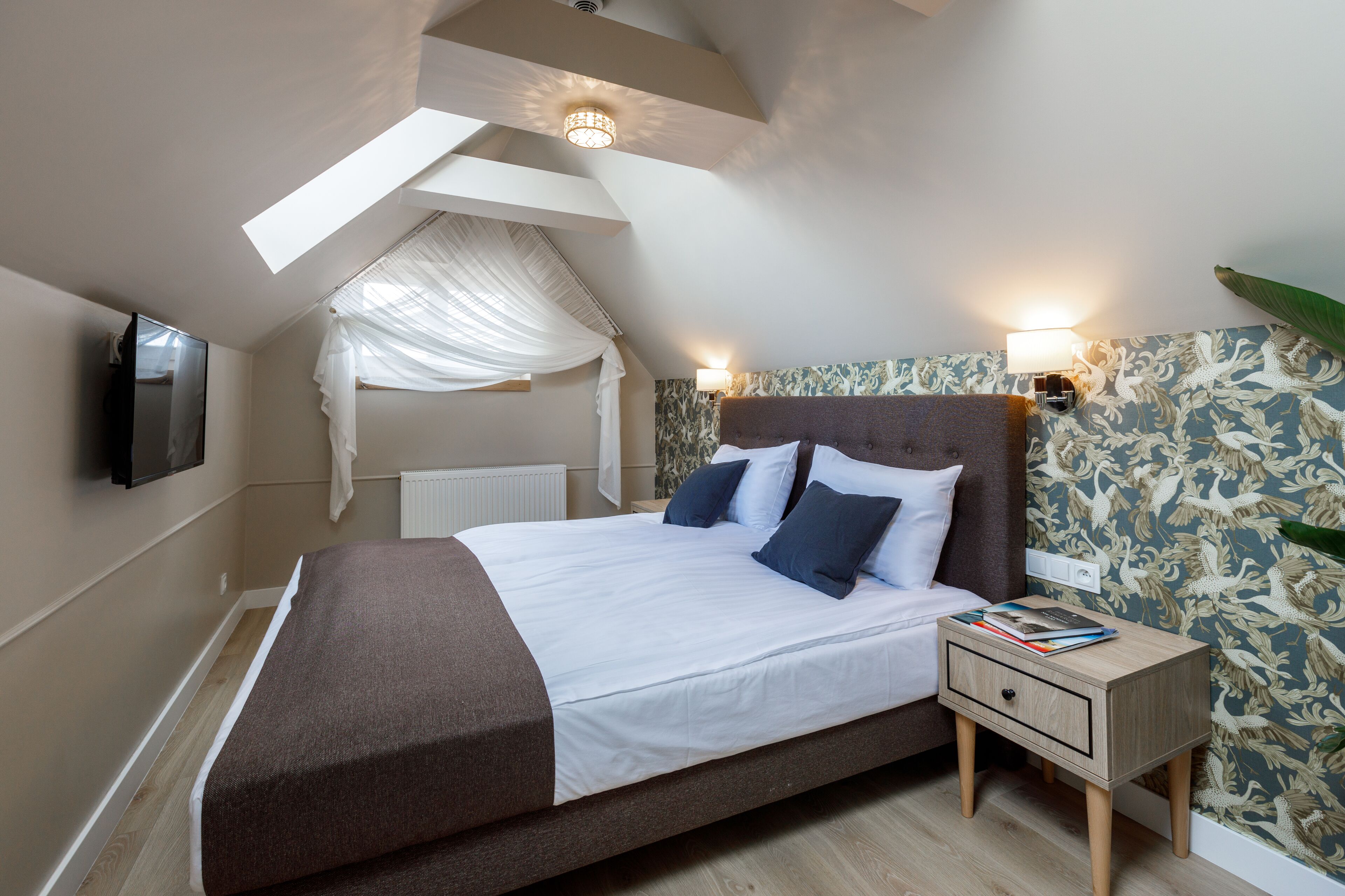 A bedroom with a large bed, a nightstand, a TV mounted on the wall, and a window with a sheer curtain.