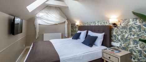 A bedroom with a large bed, a nightstand, a TV mounted on the wall, and a window with a sheer curtain.