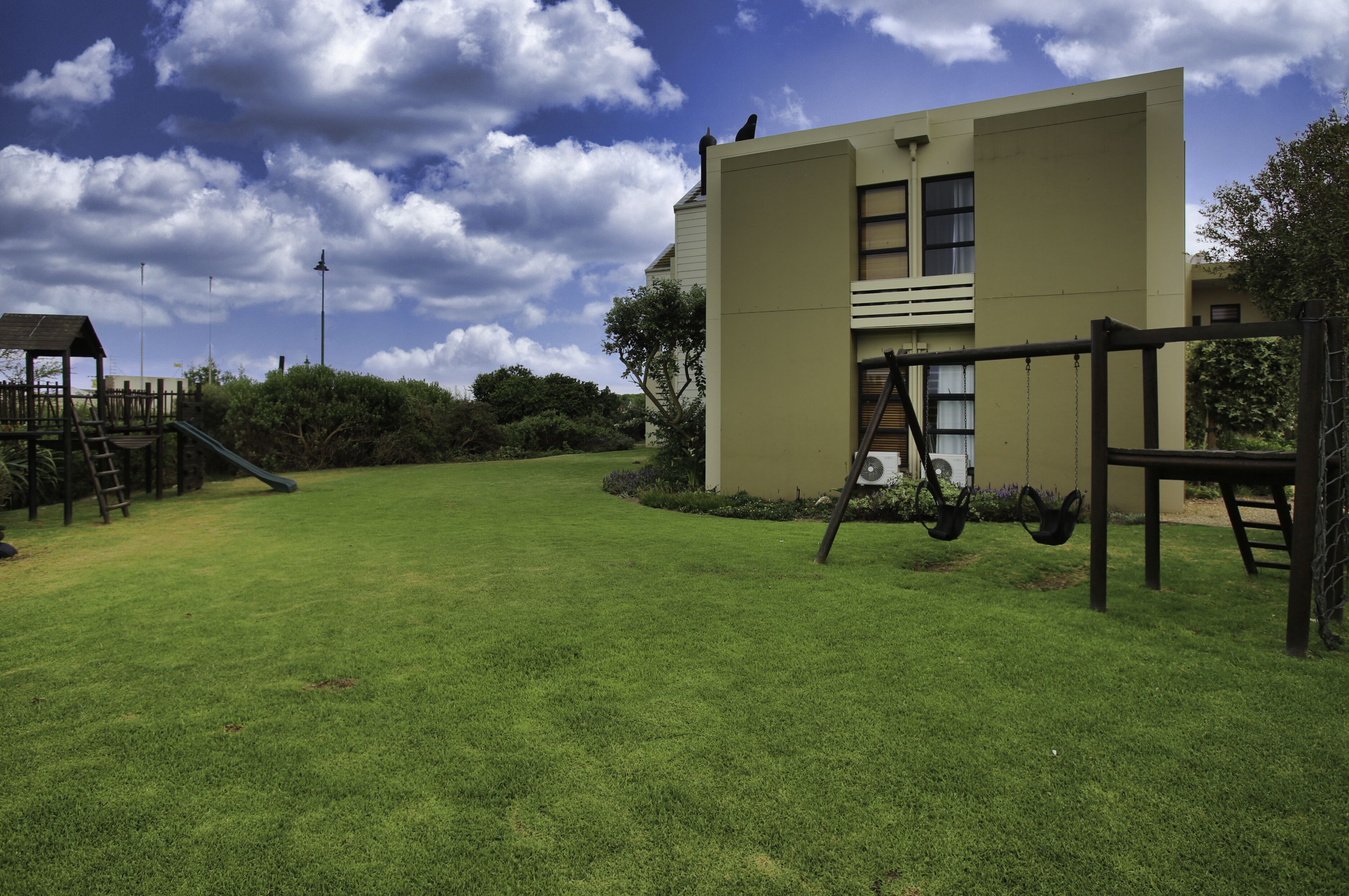 A playground with a slide and swings, a wooden playhouse, and a building with a beige exterior.